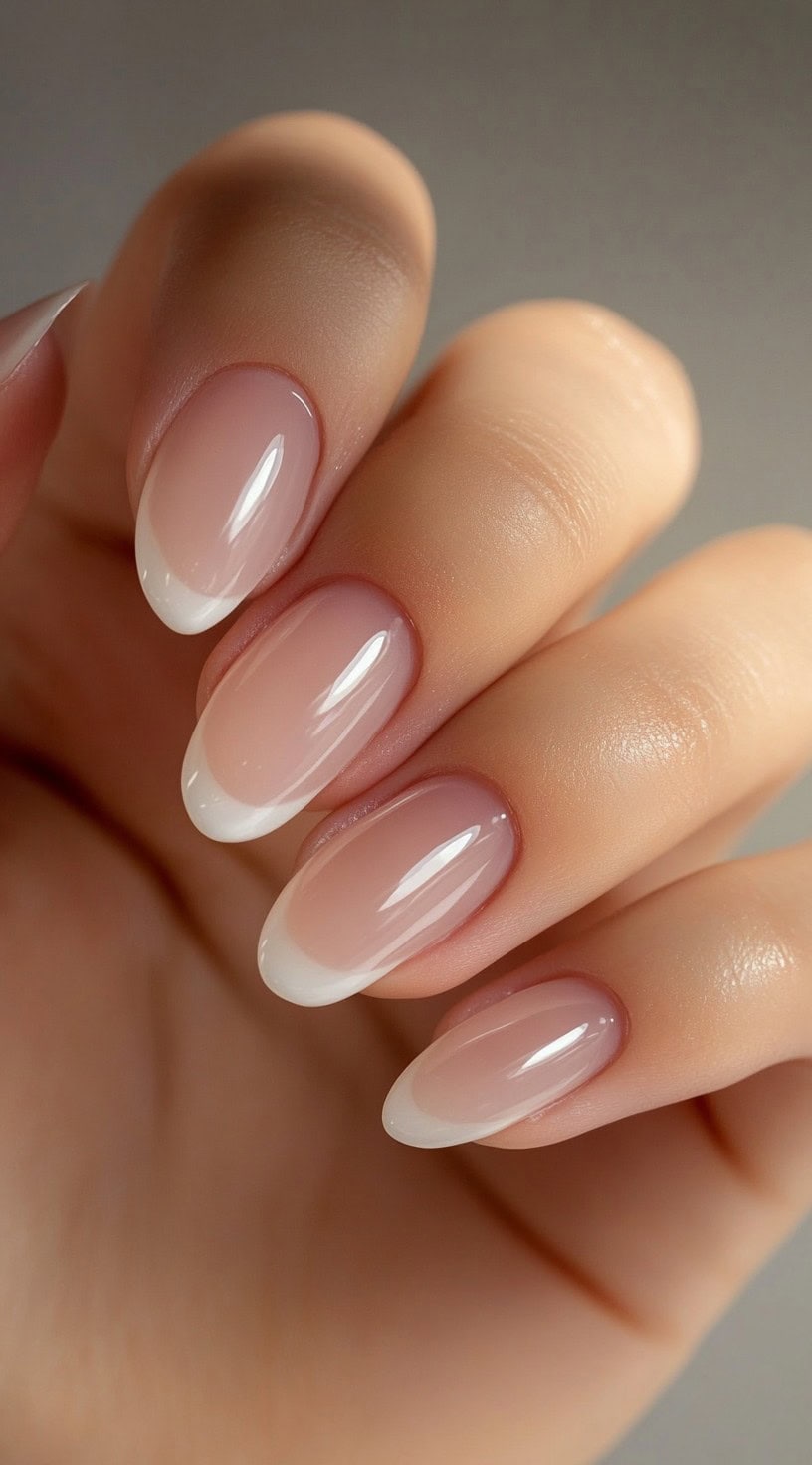 A close-up of a bride's hand with soft almond-shaped nails featuring a smooth French ombré effect, transitioning from sheer pink at the base to soft white tips.