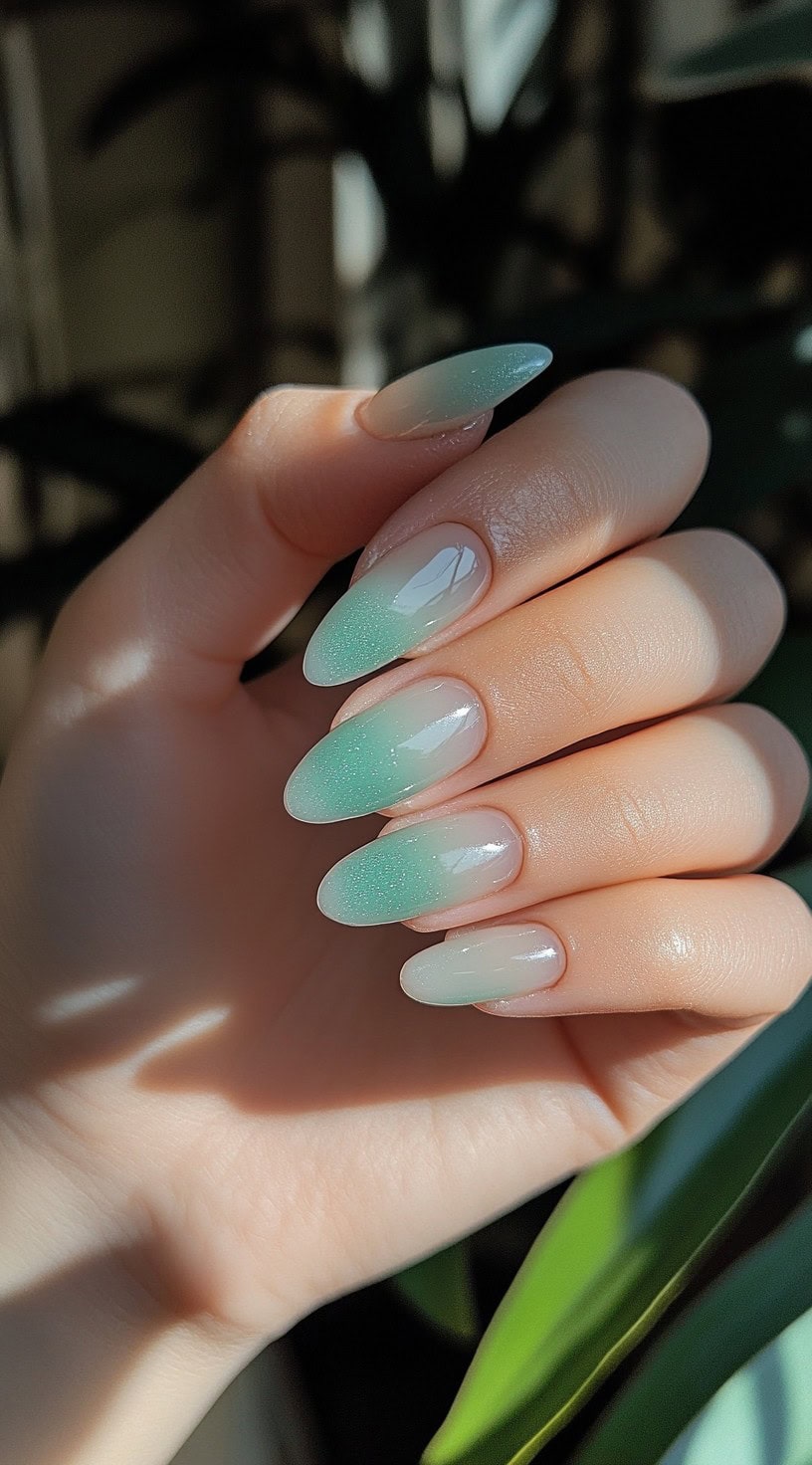 A hand with almond-shaped nails featuring a soft green ombre fade against a blurred background with natural light.
