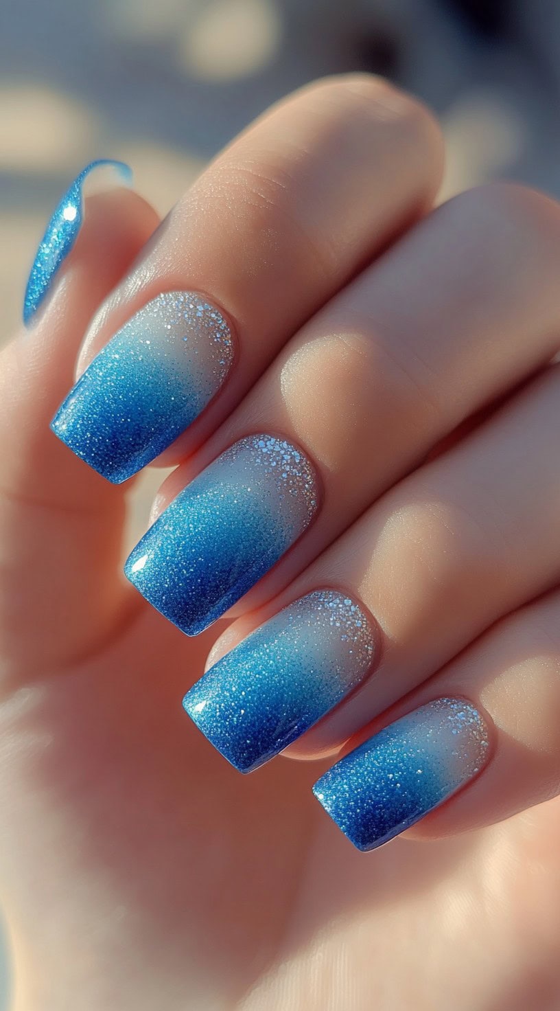 A hand with square nails fading from a soft translucent base into icy blue glitter tips.