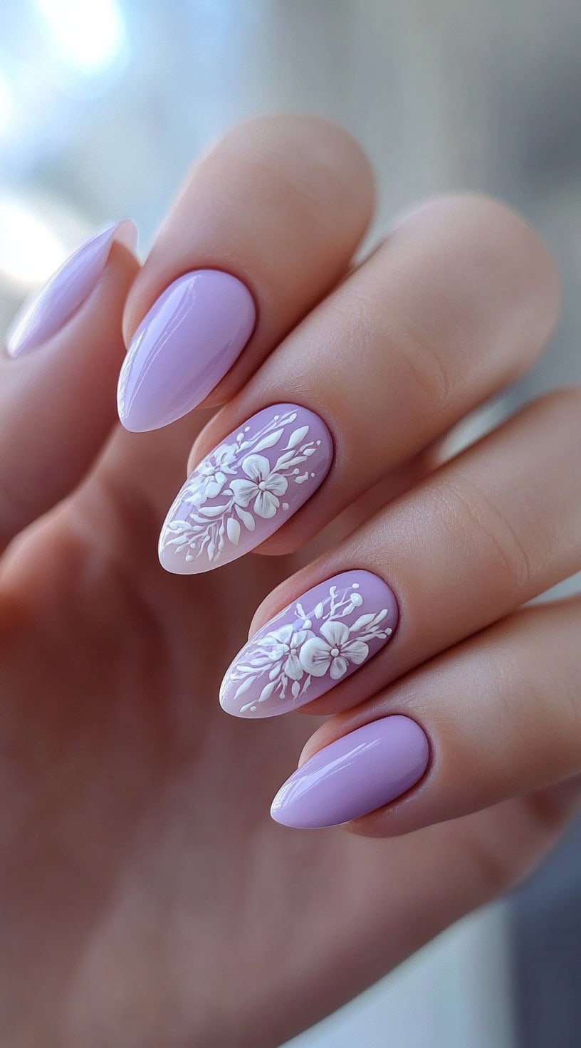 A close-up of a hand displaying soft lavender nails, with two accent nails featuring intricate 3D white floral designs.