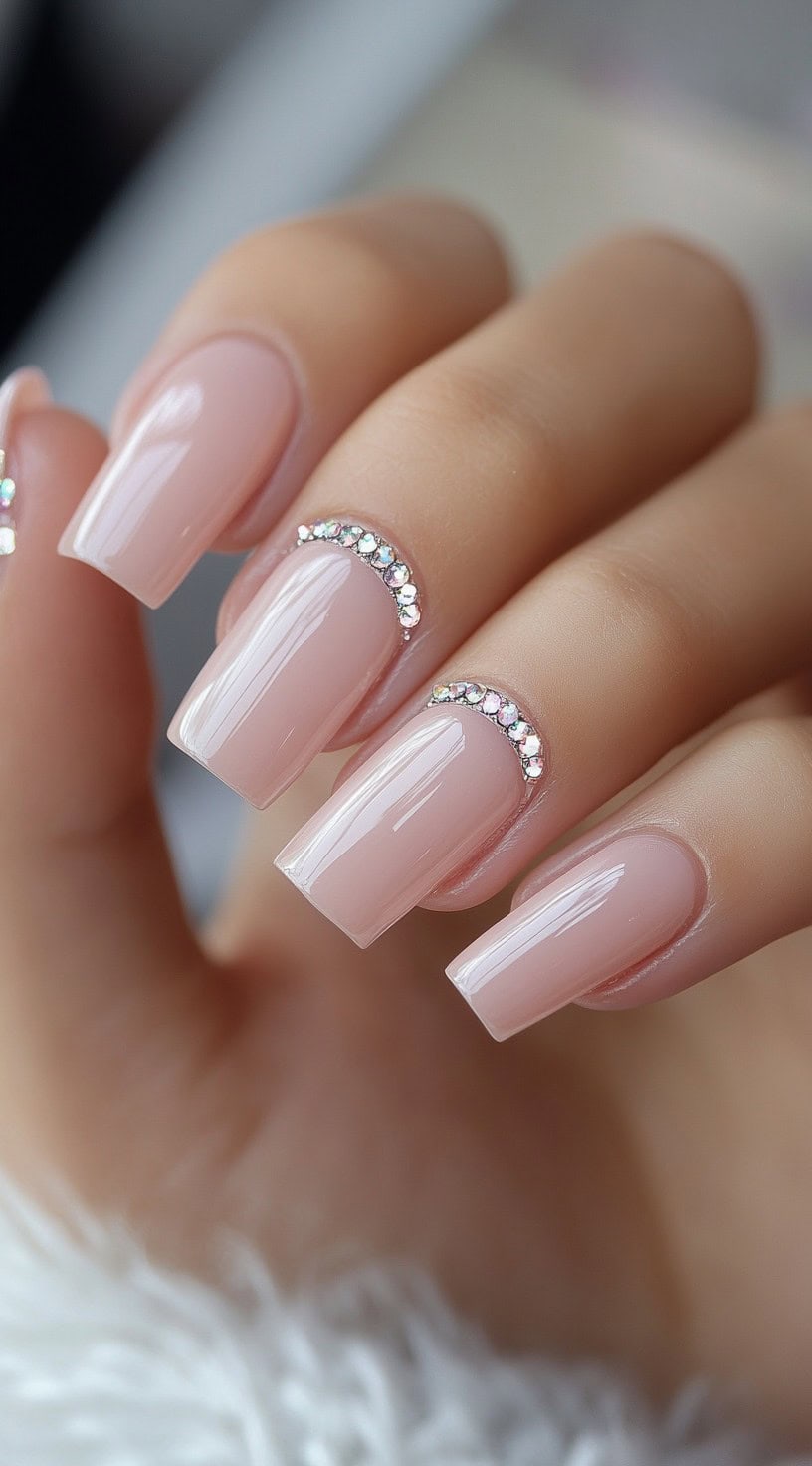 A close-up of long, coffin-shaped nails in a soft nude shade, featuring a curved row of sparkling rhinestones near the cuticle.