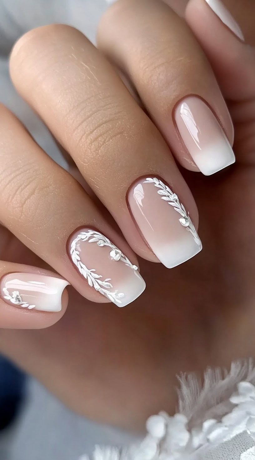 A close-up of a hand showcasing soft ombré square nails with white vine detailing and small crystal embellishments.