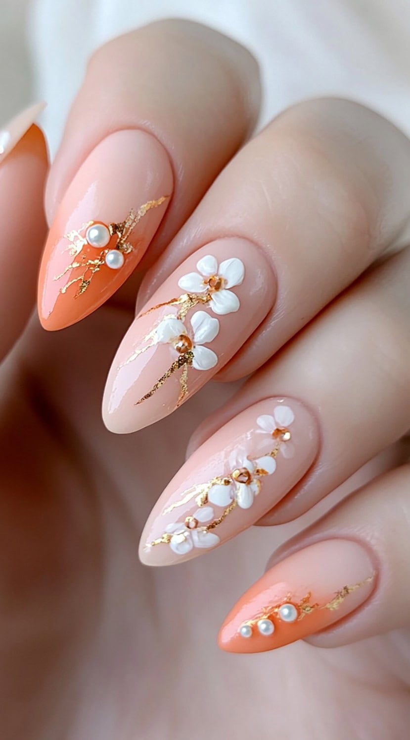 Almond nails in soft peach and nude tones, detailed with 3D white flowers, gold foil branches, and pearl accents.