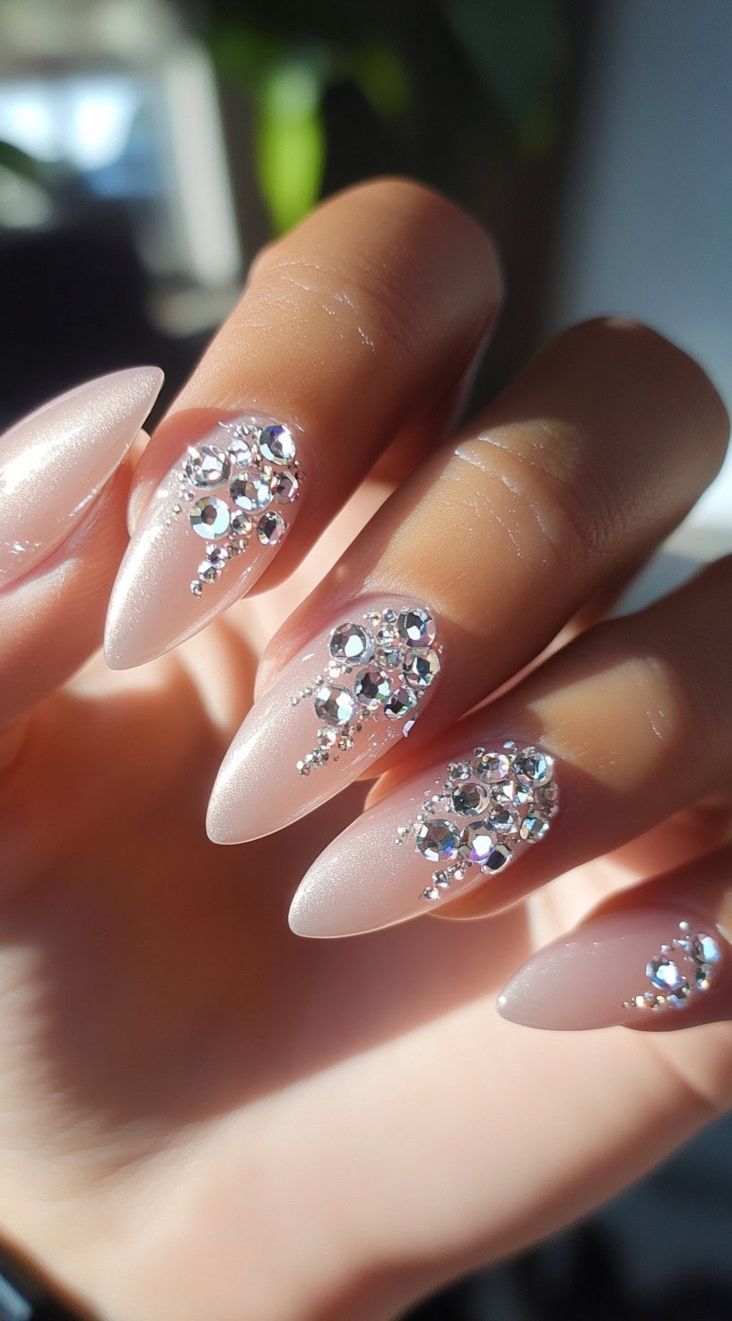 A close-up of soft pink almond-shaped nails adorned with cascading clear rhinestones, reflecting light beautifully.