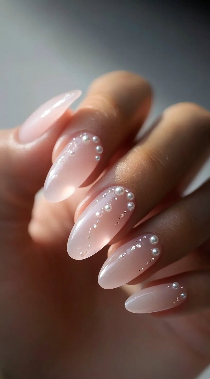 A close-up of almond-shaped nails in a soft pink shade, adorned with delicate pearls and tiny crystals arranged in a curved pattern.