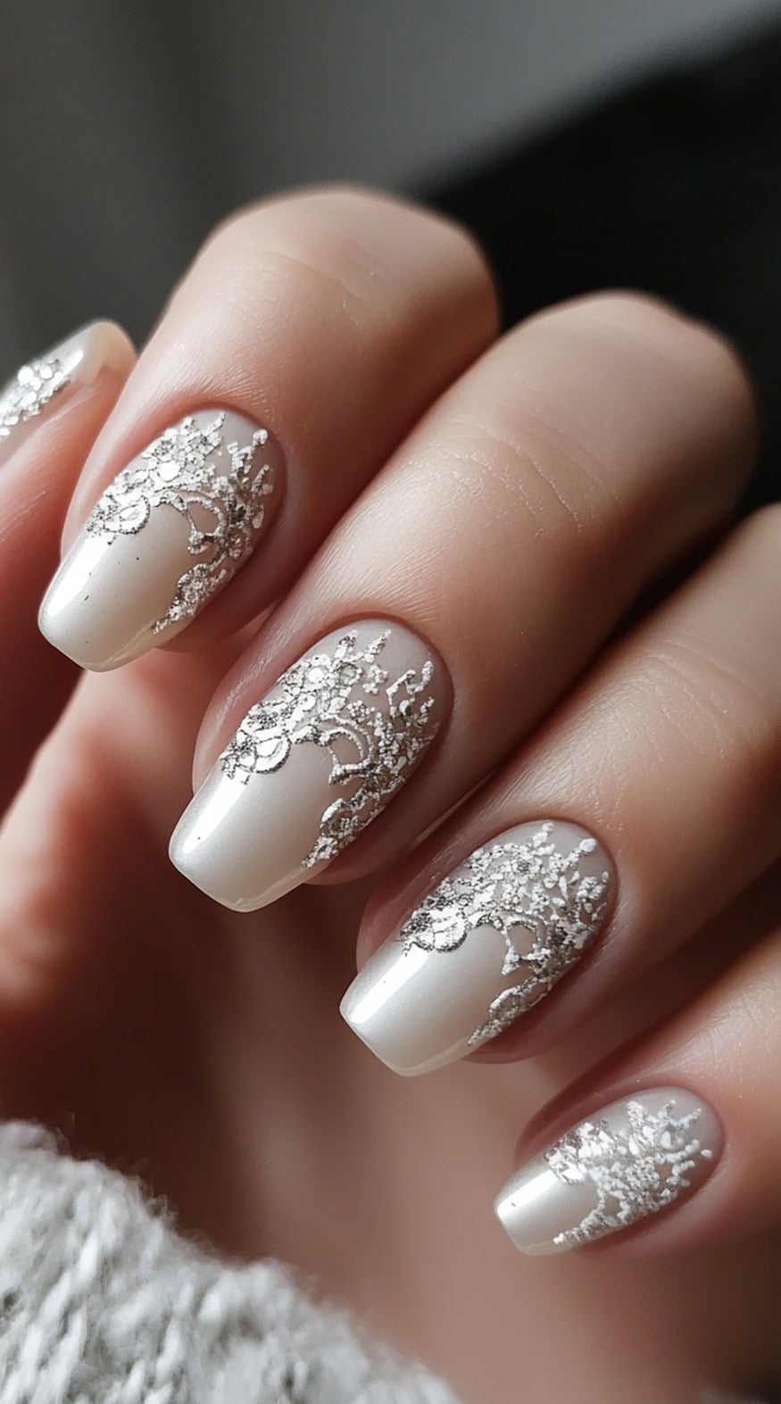 A close-up of medium-length nails with a sheer nude base, delicately decorated with intricate silver lace-like patterns.