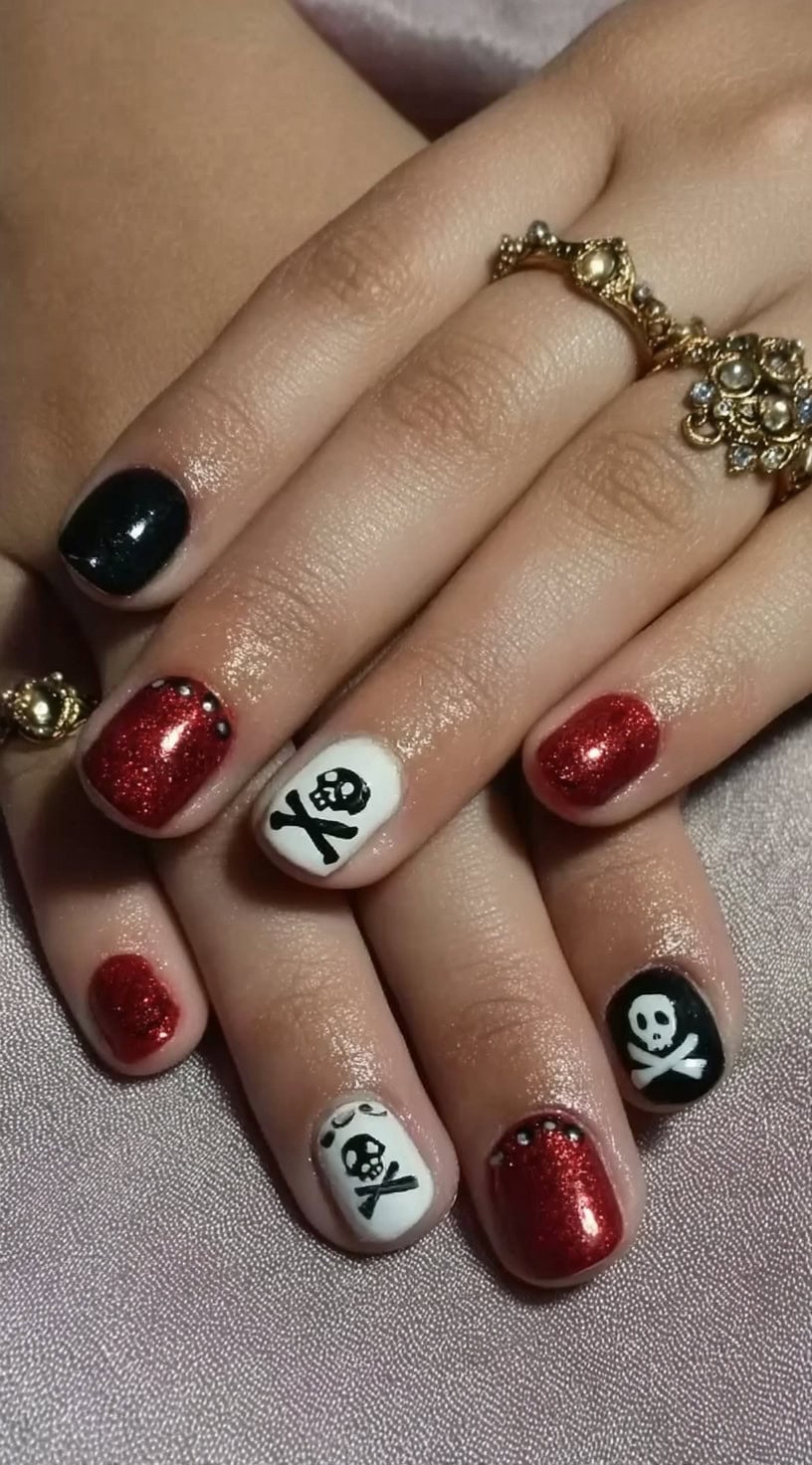 Short glittery red, white, and black nails feature skull-and-crossbones art with rhinestone accents.