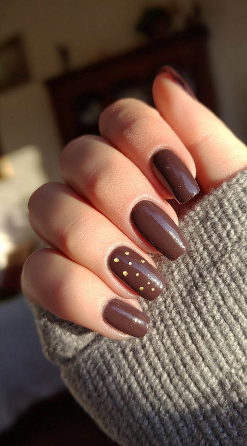 Square-shaped nails in chocolate brown polish with one accent nail decorated with vertical gold dots.