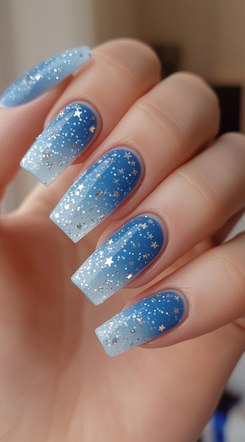 A hand with medium-length square nails featuring a blue to white ombre with star-shaped glitter accents.