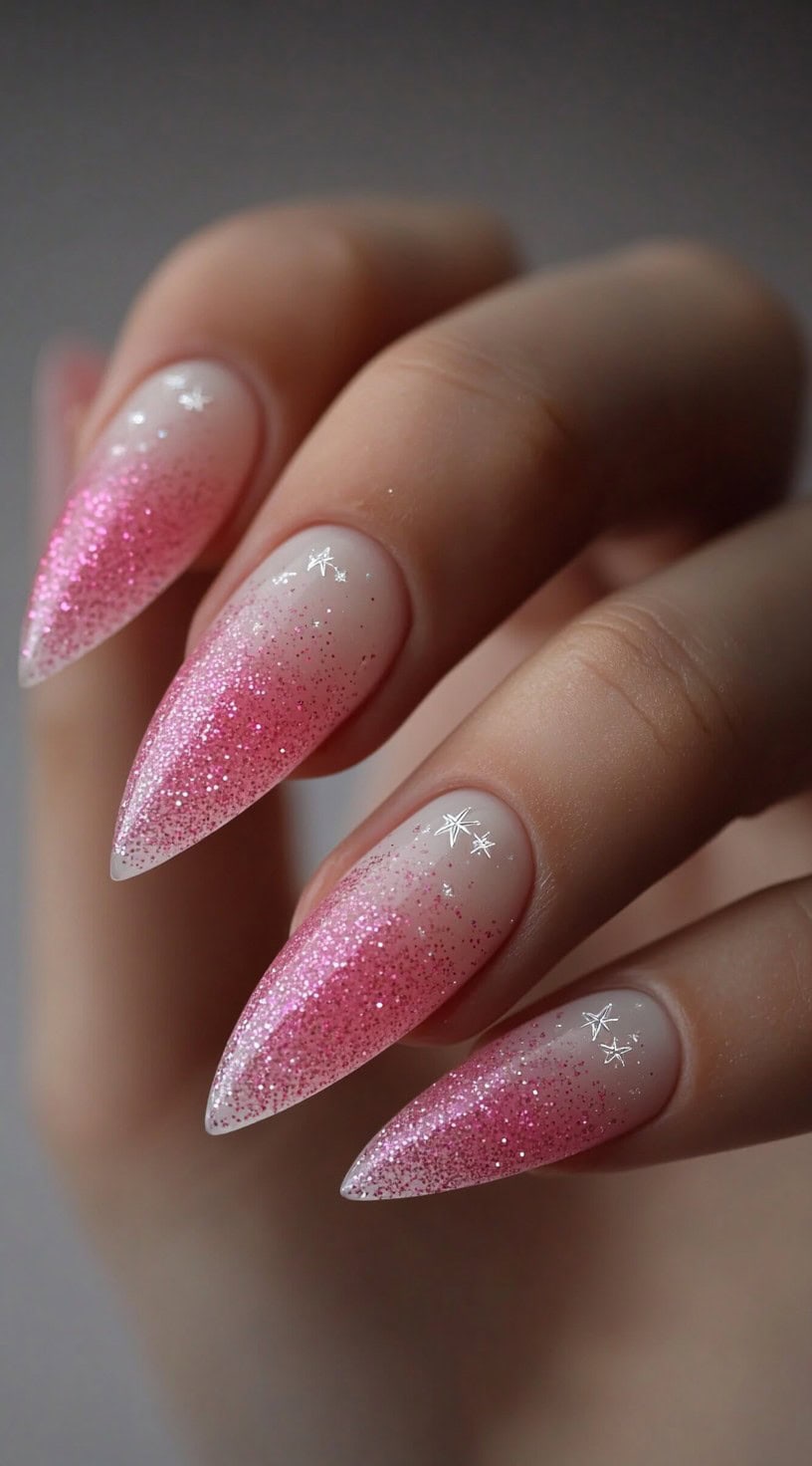 A hand with long stiletto nails featuring a gradient of bright pink glitter fading into soft nude with small white star decals.