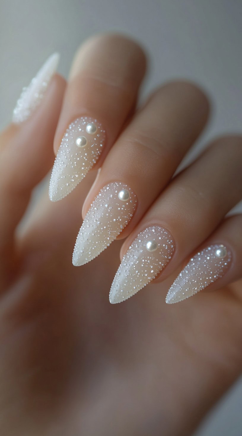 A close-up of stiletto nails with a sheer white base, textured sugar-like glitter, and pearl accents.