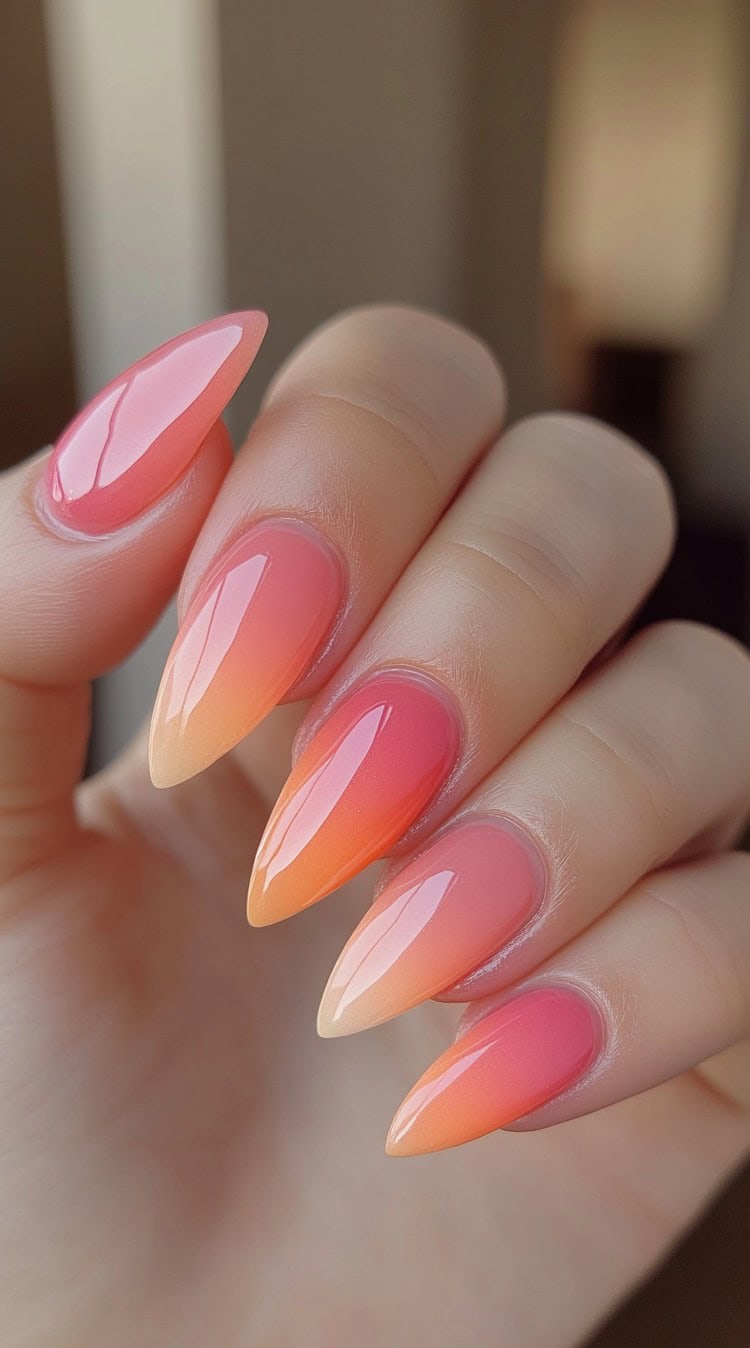 A set of long stiletto-shaped nails transitioning from a soft pink base to a warm orange gradient with a glossy finish.