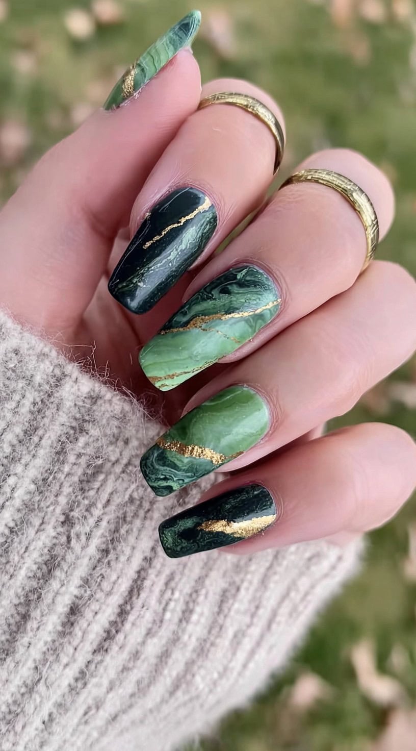 A hand with long square nails in dark and light jade swirls, each nail marked by a bold metallic gold vein.