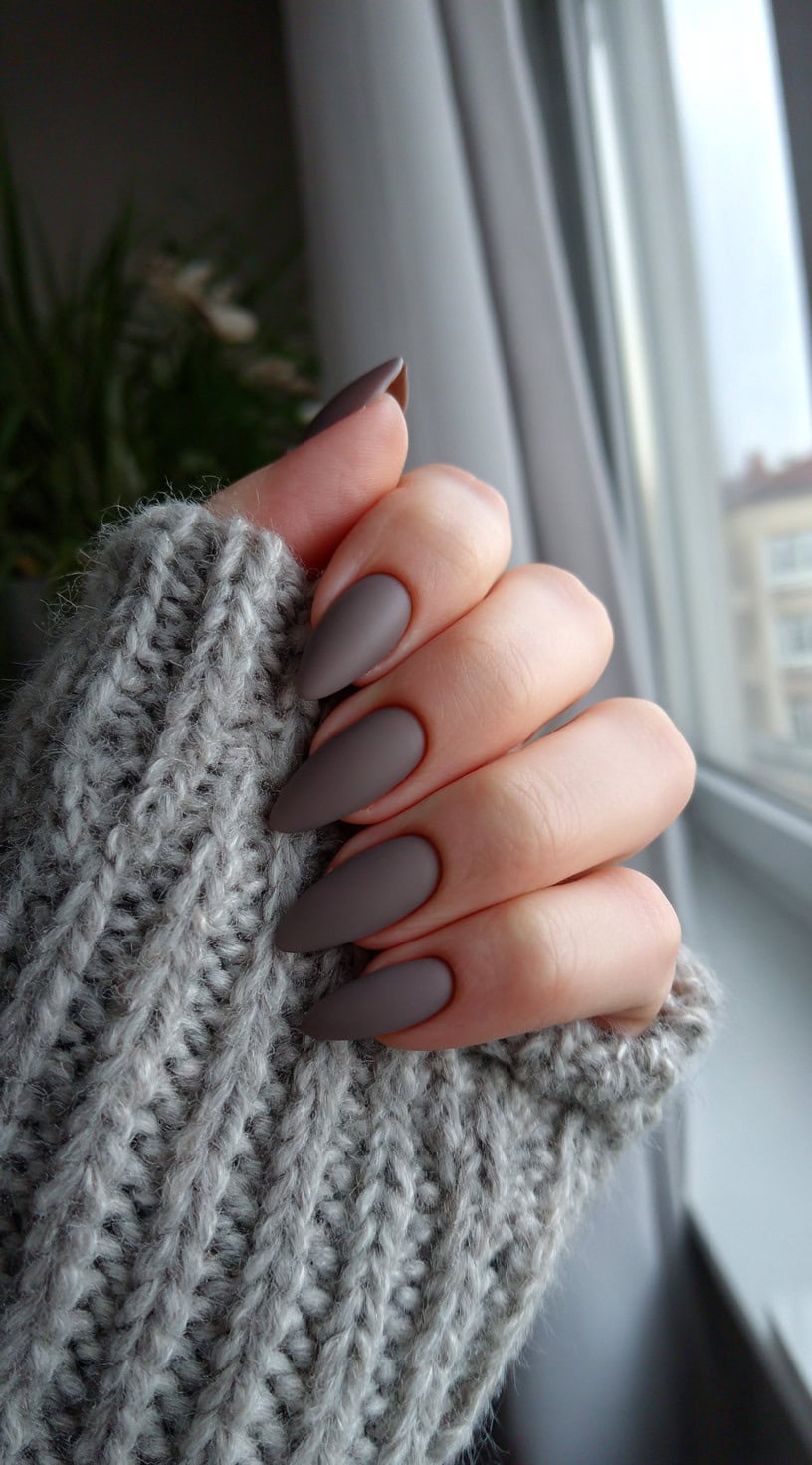 A hand with long taupe matte stiletto nails held up against a window, wrapped in a chunky gray knit sweater.