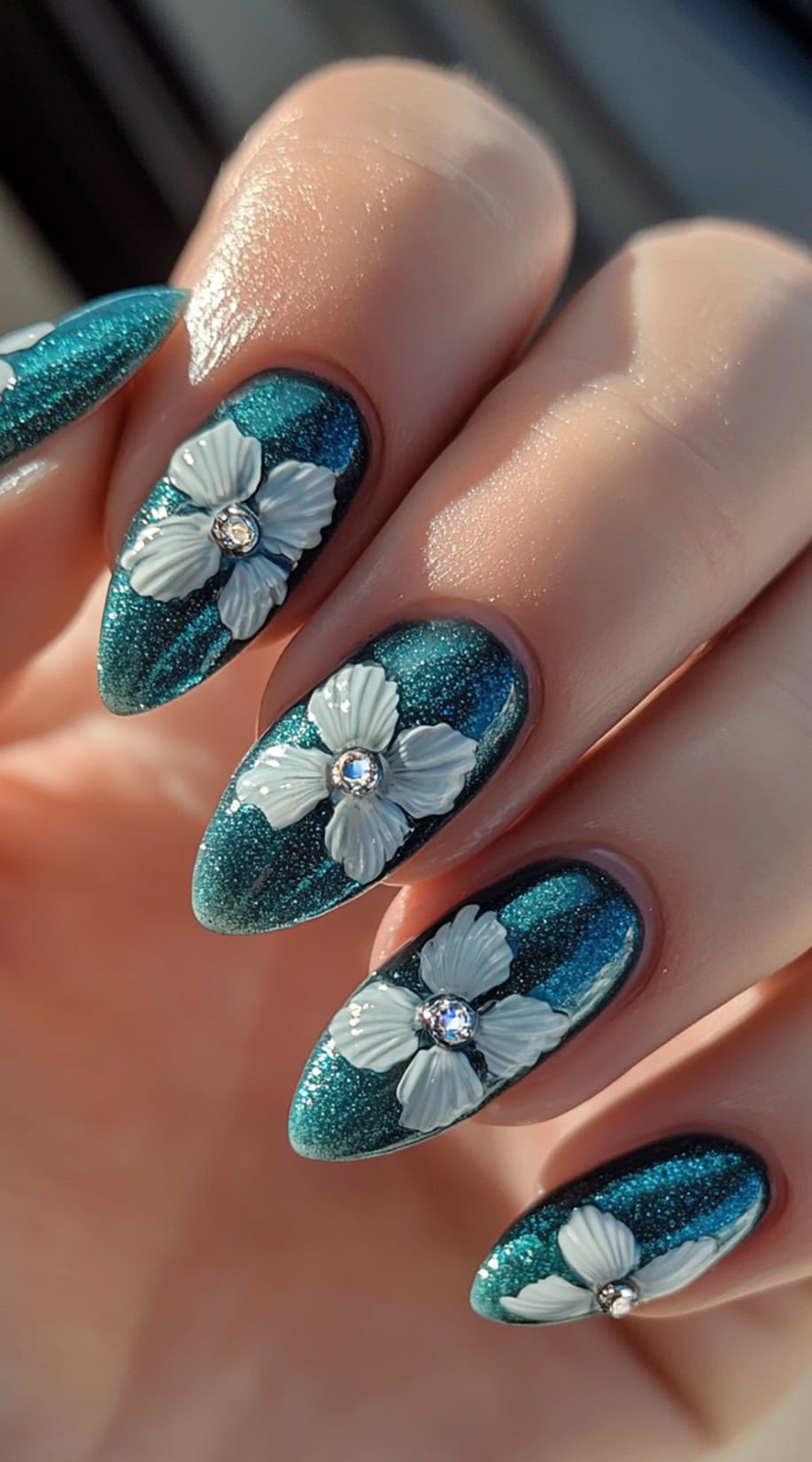 A close-up of teal glittery nails with intricate 3D white flowers and rhinestone centers.