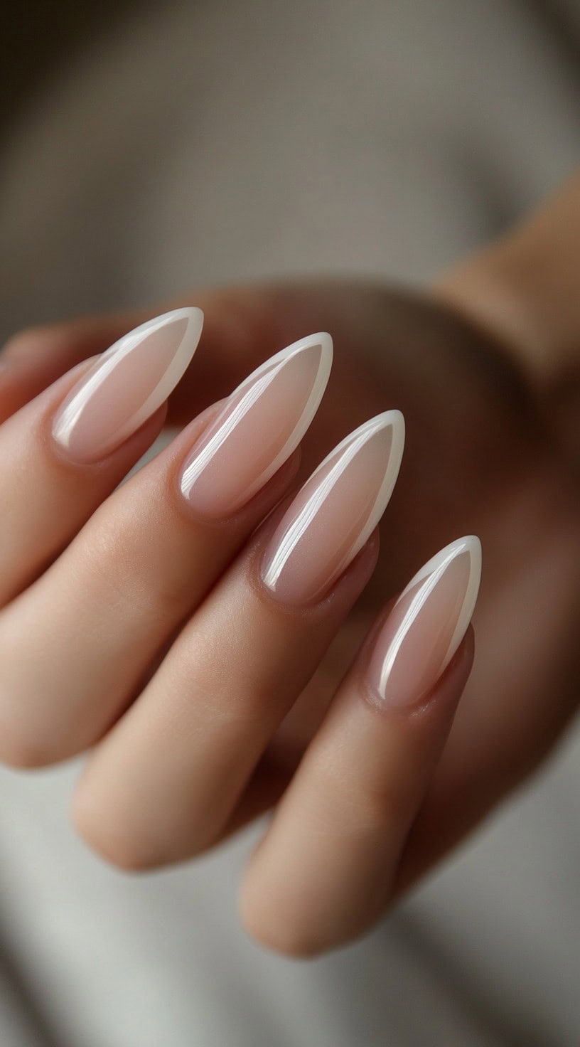 A close-up of a hand with almond-shaped nails featuring a thin white French tip on a sheer nude base.