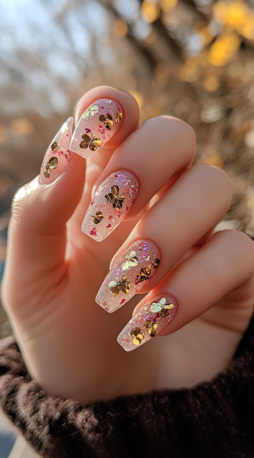 Long, coffin-shaped nails with a translucent pink base, shimmering glitter, and delicate gold butterfly embellishments for a whimsical effect.