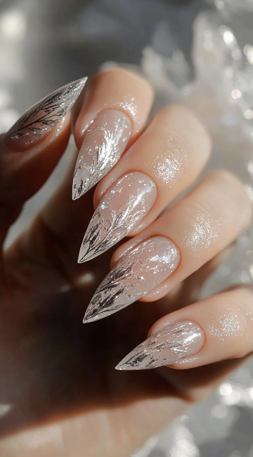 A close-up of long, transparent stiletto nails with delicate silver foil details, creating a shimmering, icy effect.