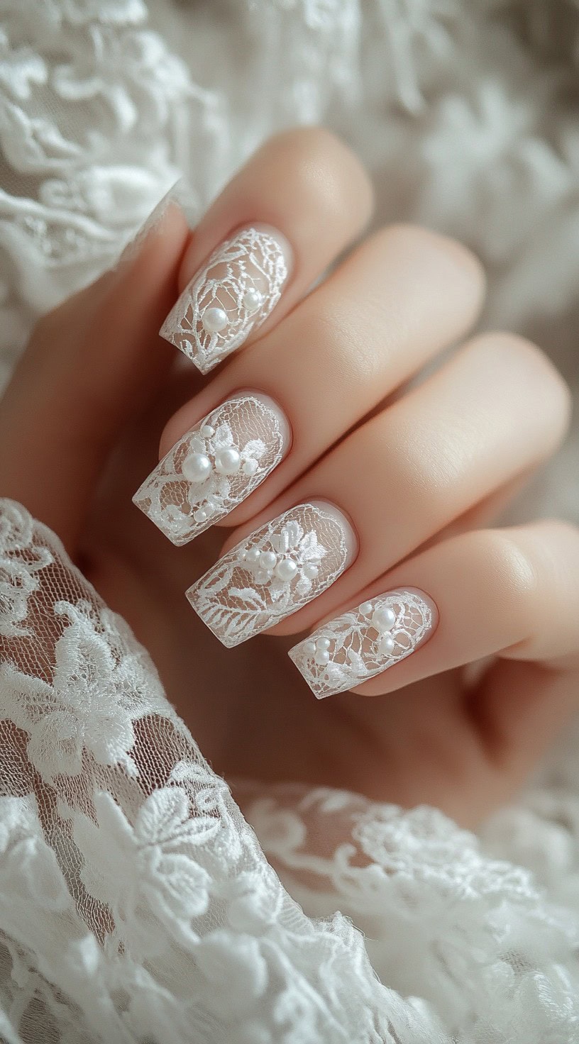 Square-shaped nails featuring intricate white lace patterns over a sheer nude base, adorned with delicate pearl accents.