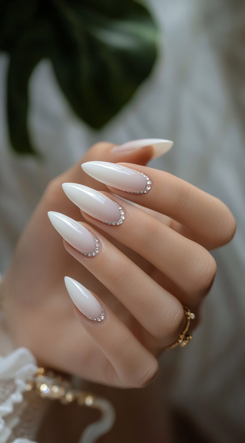 A hand with long, almond-shaped nails painted in glossy white, featuring a delicate line of small crystals along the cuticles.