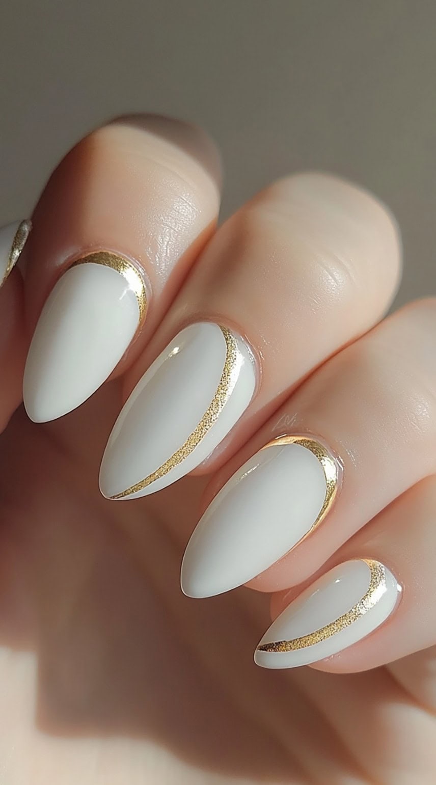 A hand with almond-shaped nails painted in a soft white shade, featuring delicate gold arc detailing along the cuticles and edges.