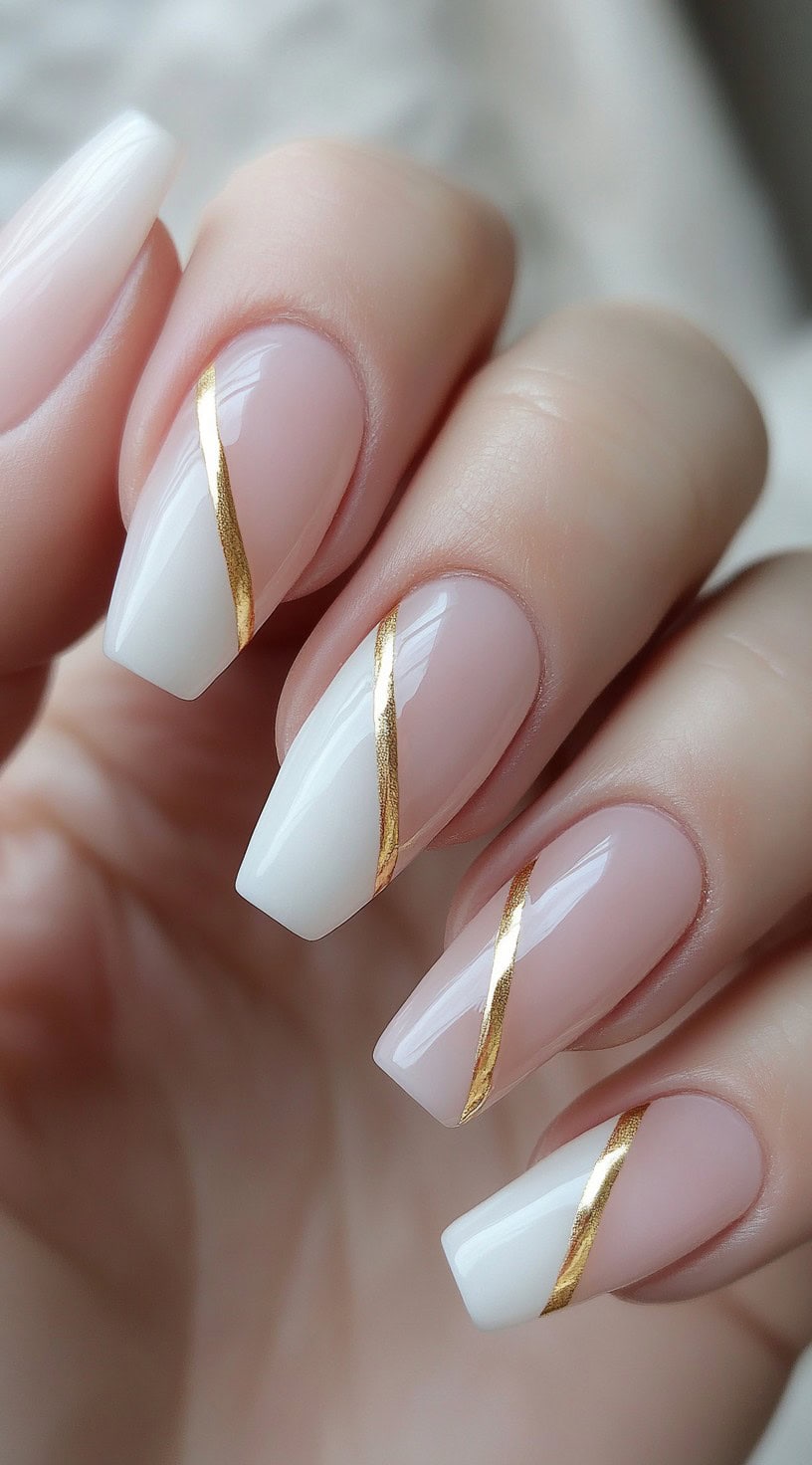 A close-up of a hand with glossy nude and white French-style diagonal nails, featuring thin gold metallic accents for an elegant and sophisticated look.