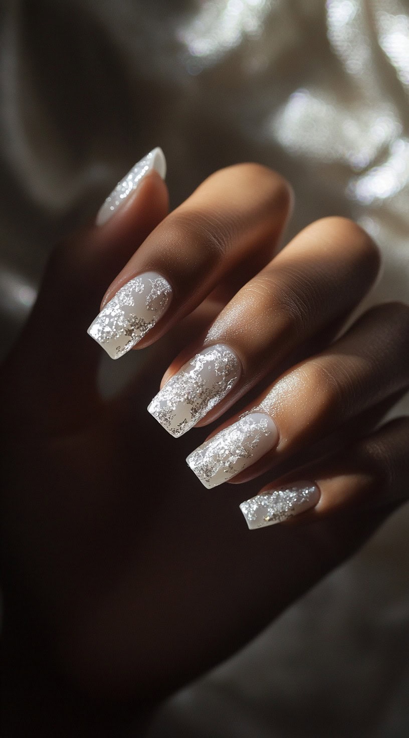 A close-up of square-shaped nails painted white with silver foil accents creating a shimmering textured effect.