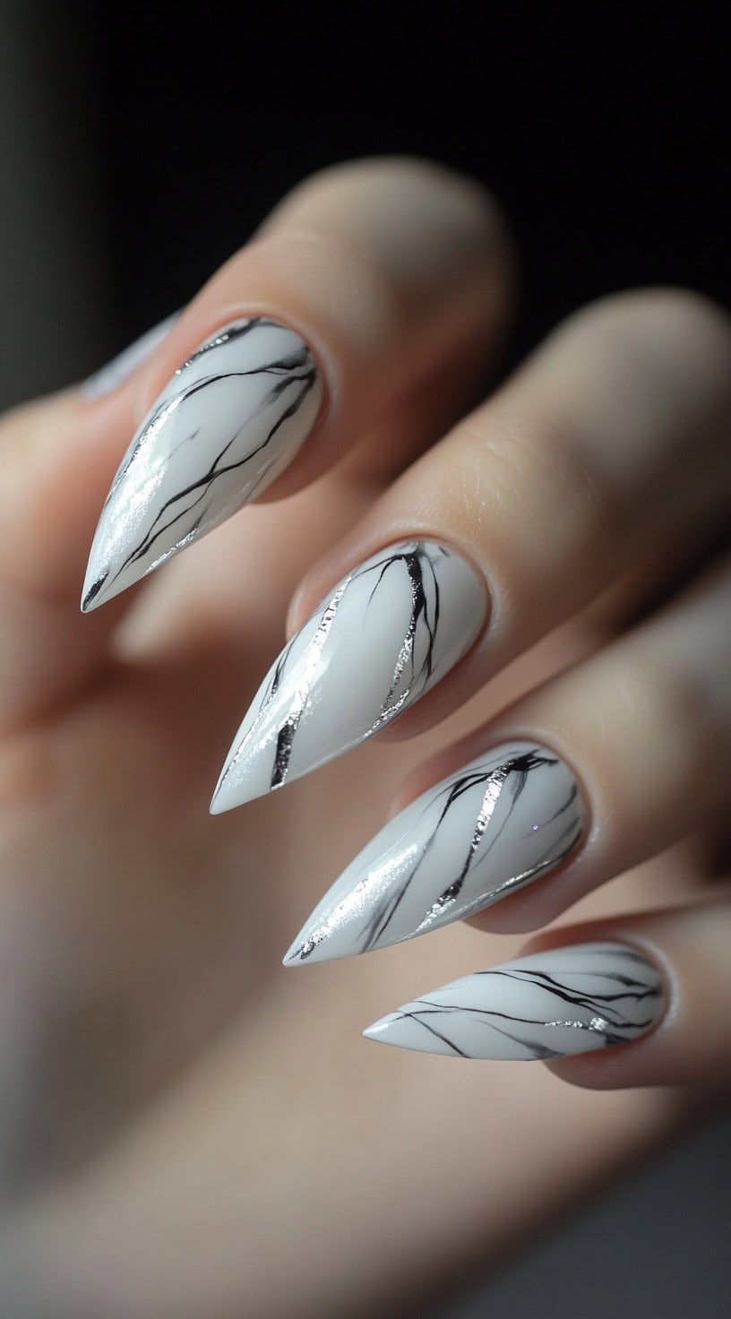 A close-up of long stiletto nails with a white marble base, accented with fine black veining and silver metallic streaks.