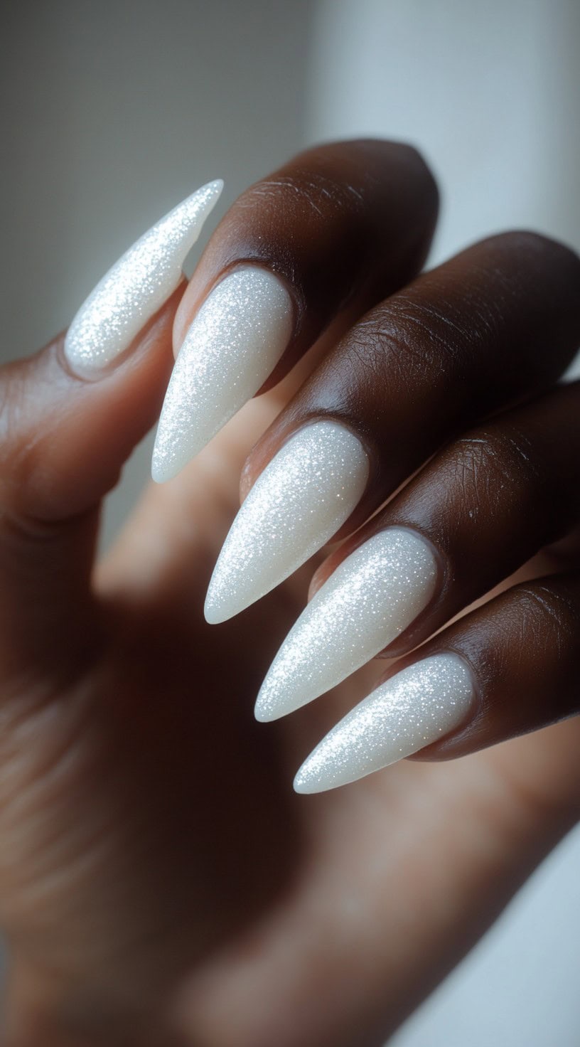 A close-up of long stiletto-shaped nails coated in sparkling white gel, creating a glamorous and eye-catching wedding look on deep-toned skin.