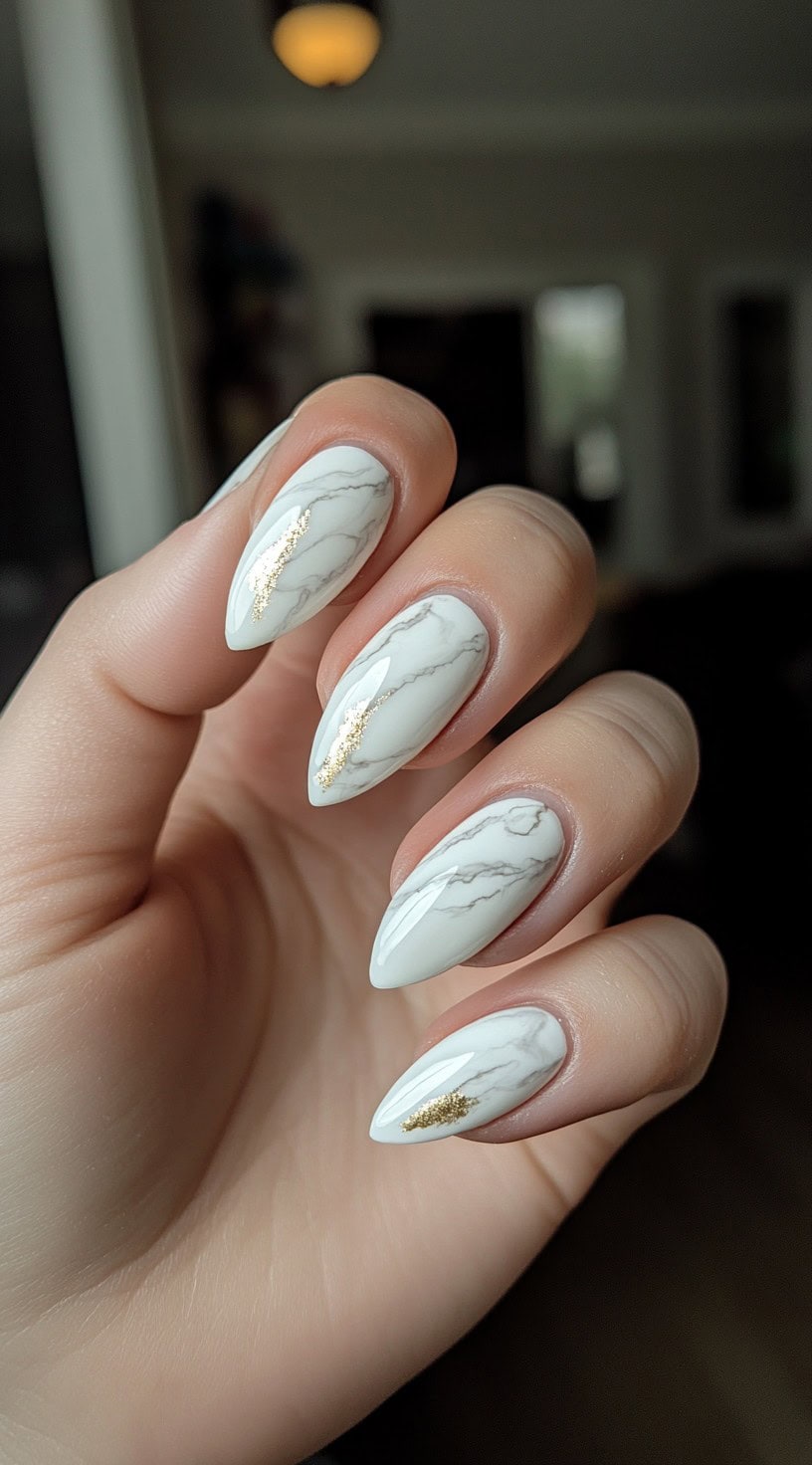 A close-up of almond-shaped nails featuring a white marble design with thin gold foil accents.