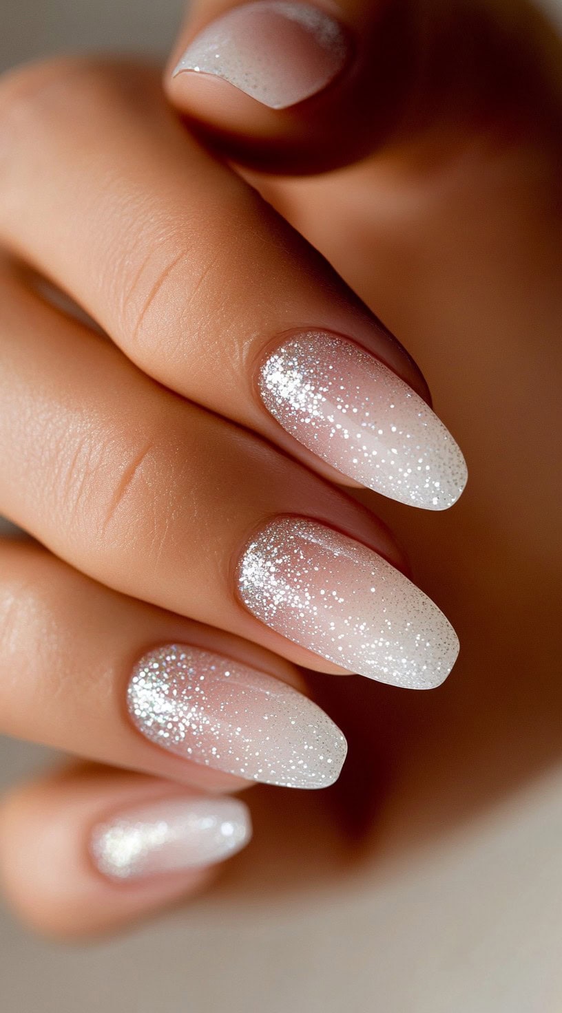 A hand featuring almond-shaped nails with a white-to-pink ombré gradient and silver glitter dusted at the tips.