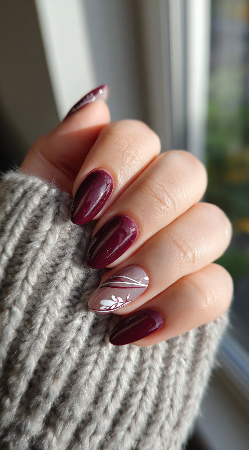 A hand showing burgundy almond nails with one accent nail featuring soft taupe polish and a delicate white leaf design.