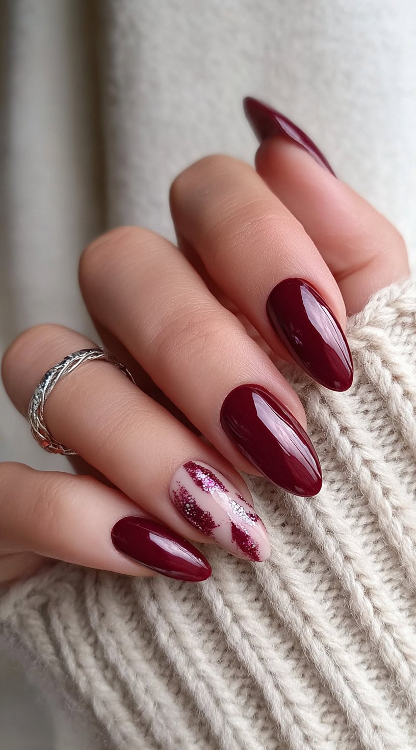 A hand with almond-shaped nails painted in deep wine, with one accent nail featuring a nude base and angled burgundy glitter brushstrokes.